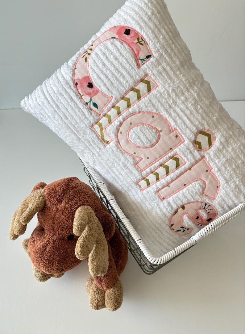 May include: White plush pillow with the name "Claire" embroidered in pink letters with a floral and chevron pattern. The pillow is in a wire basket with a brown plush moose toy.