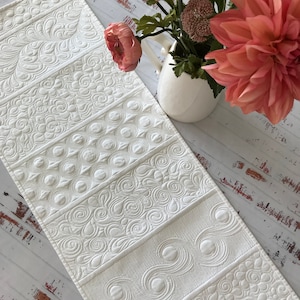 May include: White quilted table runner with a variety of embossed patterns. The runner is made of a soft, white fabric and has a subtle sheen.