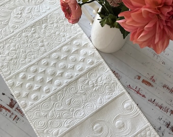 May include: White quilted table runner with a variety of embossed patterns. The runner is made of a soft, white fabric and has a subtle sheen.