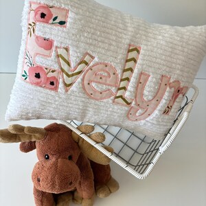 May include: White plush pillow with the name "Evelyn" embroidered in pink and gold fabric with a floral pattern. The pillow is in a wire basket with a brown plush moose toy.