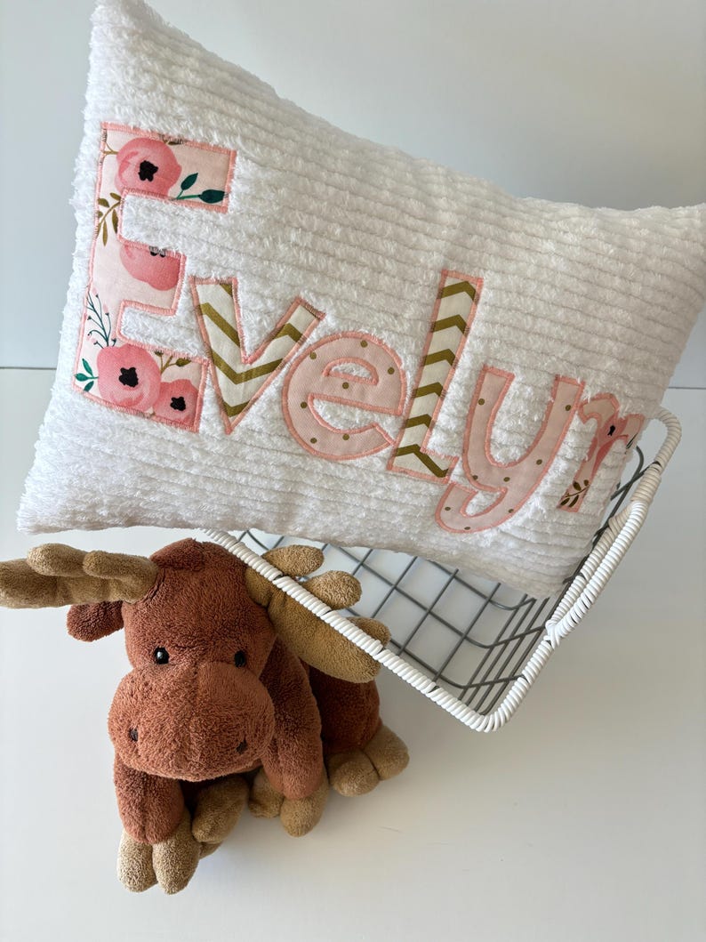 May include: White plush pillow with the name "Evelyn" embroidered in pink and gold fabric with a floral pattern. The pillow is in a wire basket with a brown plush moose toy.