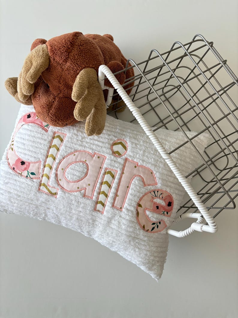 May include: White pillow with the name "Claire" embroidered in pink letters with floral and gold accents. A brown plush moose toy and a wire basket are in the background.