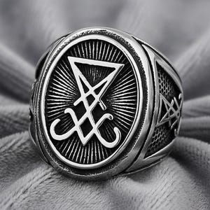 Puede incluir: Un anillo plateado con una cara ovalada que presenta un símbolo estilizado dentro de un diseño radiante. La banda del anillo tiene un patrón texturizado y un diseño de estrella en los lados. El anillo está sobre un fondo gris suave.