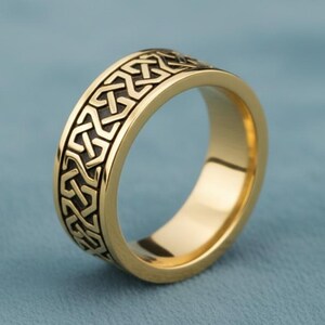 May include: A gold-colored ring with a detailed Celtic knot pattern around the band. The ring is made of a shiny, polished metal and is set against a blue background. The ring's design features interwoven lines and shapes.