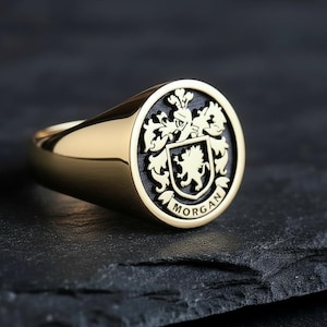 May include: A gold signet ring with a black and gold crest. The crest features a shield with a lion and floral elements, with the name "MORGAN" inscribed below. The ring is set on a dark surface.