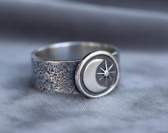 May include: A silver ring with a textured band and a circular emblem featuring a crescent moon and a starburst design. The ring has a rustic, antique appearance.
