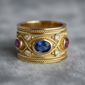 Puede incluir: Un anillo de oro ancho con un diseño intrincado. Presenta una gran gema azul ovalada en el centro, flanqueada por dos gemas rojas más pequeñas. Pequeñas piedras claras y detalles decorativos en forma de cuerda completan el diseño.