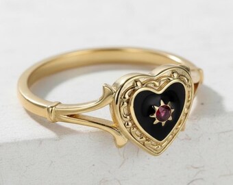 May include: A gold ring featuring a heart-shaped face. The heart is black with a small red gemstone at its centre. The ring has an ornate design around the heart and on the band's supports.