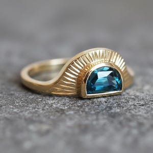 May include: A gold ring with a half-moon design, featuring a vibrant blue gemstone. The ring's band has a textured, sunburst-like pattern around the gemstone. The ring is set against a gray surface, highlighting its intricate details.