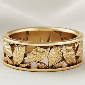 Op de afbeelding: Een goudkleurige ring met een gedetailleerd bladerenontwerp. De ring heeft een band van bladeren die rond de omtrek is gesneden, waardoor een opengewerkt patroon ontstaat. De bladeren hebben gedetailleerde texturen en de ring heeft een gepolijste afwerking.