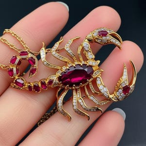 May include: A gold scorpion pendant necklace featuring a large, oval, deep red gemstone. The scorpion's body and claws are embellished with small, clear gemstones. The necklace chain also has red gemstones.