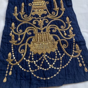 以下が含まれることがあります： 金色の刺繍シャンデリアのデザインが施された濃紺の生地。シャンデリアには、ろうそくのような要素やぶら下がっているビーズのアクセントなど、複雑なディテールが施されています。生地はキルティング模様です。