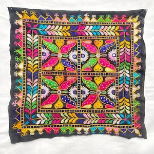 Puede incluir: Textil cuadrado negro con intrincados bordados coloridos. El diseño presenta patrones florales y geométricos en rosa, amarillo, verde y azul, delineados en dorado. Los bordes también están bordados con un patrón floral repetitivo.