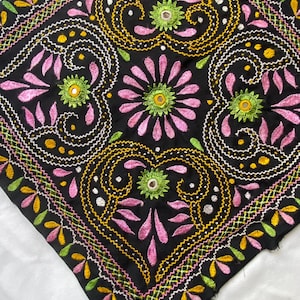 May include: Black square textile with intricate embroidery. The design features pink, yellow, green, and white floral and geometric patterns. The embroidery is detailed, with a mix of stitches creating a textured appearance. The textile is likely a decorative item.