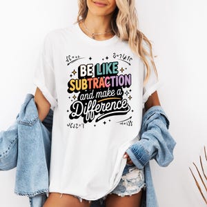May include: White t-shirt with the phrase "BE LIKE SUBTRACTION and make a Difference" in a colorful, retro font. The shirt has a crew neck and short sleeves. The design includes mathematical symbols and star accents.