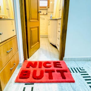Può includere: Un tappetino da bagno rettangolare rosso con le parole "NICE BUTT" in lettere rosse in grassetto. Il tappetino è posto davanti a una porta, con uno scorcio di un interno bagno visibile sullo sfondo.
