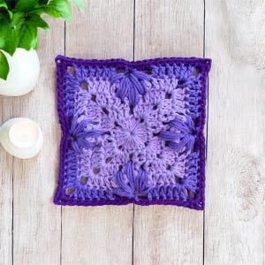 Peut inclure: Un élément carré fait à la main au crochet, dans des tons de violet. Le centre présente un motif floral, avec des éléments en forme de pétales en relief. La bordure est violet foncé, créant un bord défini. Il s'agit probablement d'un objet décoratif ou d'une partie d'un projet artisanal plus vaste.