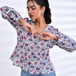 Puede incluir: Una blusa blanca con estampado floral en rojo, morado y verde. La blusa con cuello en V tiene mangas largas con puños abotonados. El diseño floral se repite en la tela. La blusa se usa con jeans azules.