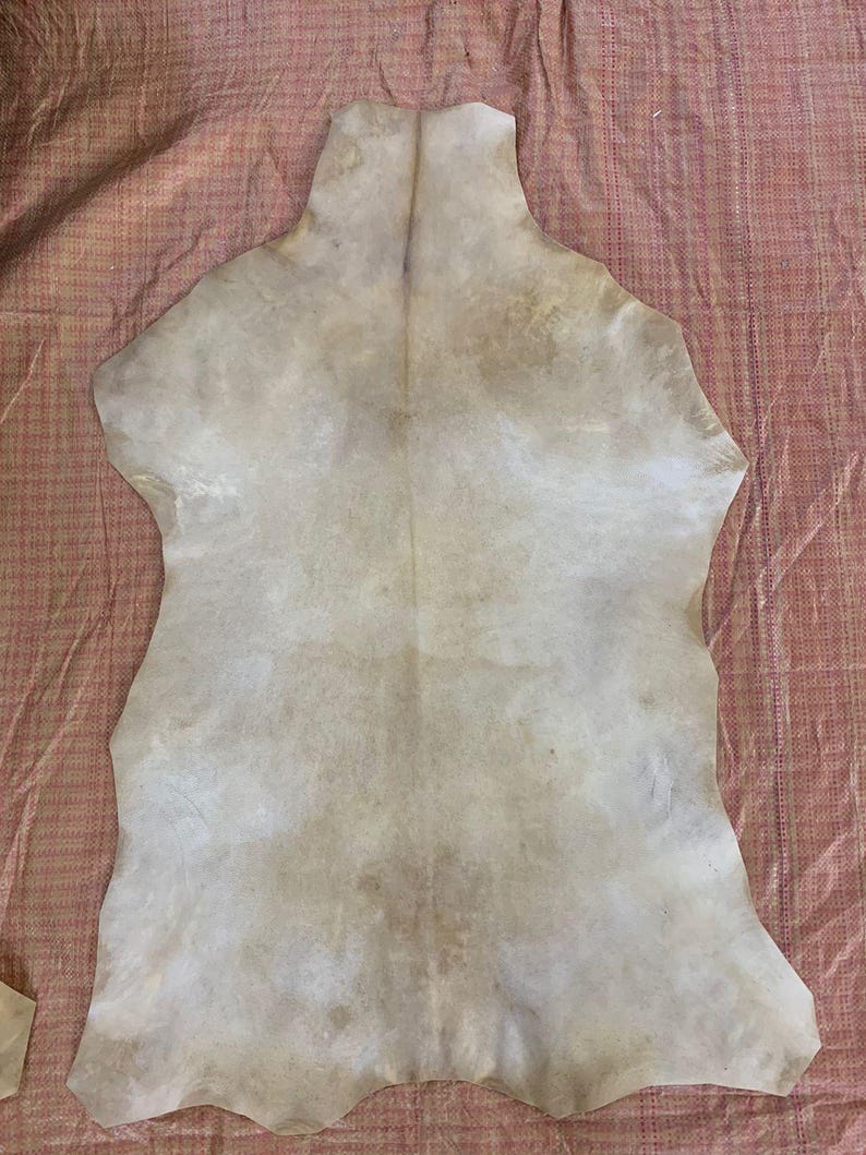 May include: A large, irregularly shaped piece of tanned leather, primarily off-white with subtle brown and tan markings. The hide is laid out on a textured, reddish-pink fabric, showcasing its natural form and variations in color and texture.