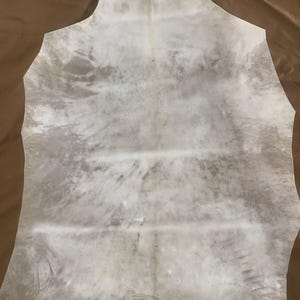 May include: A large, irregularly shaped piece of tanned leather, primarily off-white with mottled gray and brown markings. The hide has a natural, organic appearance, with visible grain and texture. The edges are roughly cut, suggesting it's ready for crafting or design projects.
