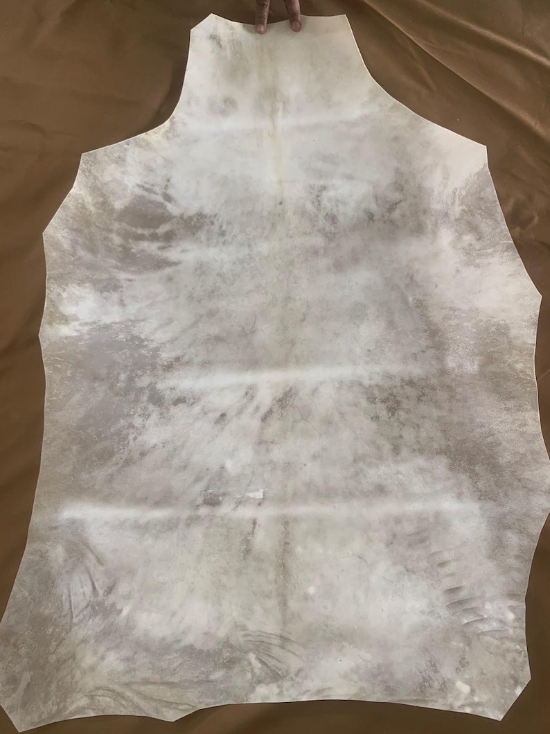 May include: A large, irregularly shaped piece of tanned leather, primarily off-white with mottled gray and brown markings. The hide has a natural, organic appearance, with visible grain and texture. The edges are roughly cut, suggesting it's ready for crafting or design projects.