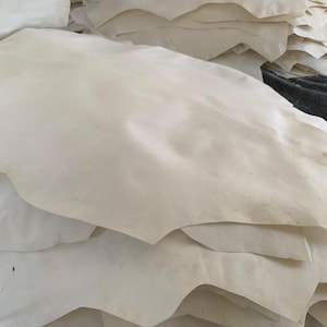 May include: A stack of off-white leather hides. The hides have irregular edges and a slightly textured surface. The leather appears to be natural and undyed, with subtle variations in color.
