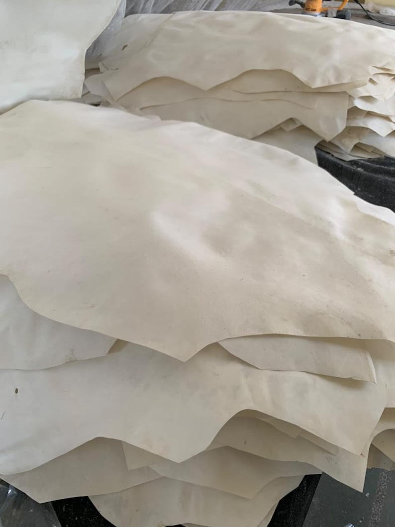 May include: A stack of off-white leather hides. The hides have irregular edges and a slightly textured surface. The leather appears to be natural and undyed, with subtle variations in color.