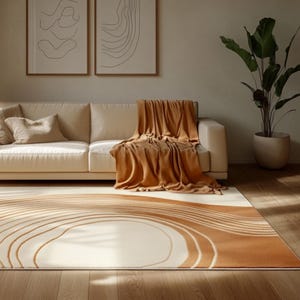 Pode incluir: Um salão moderno com um sofá creme, um cobertor castanho e almofadas. Um grande tapete com fundo creme e design de ondas castanhas domina o chão. Duas obras de arte abstratas emolduradas adornam a parede, criando um ambiente elegante.