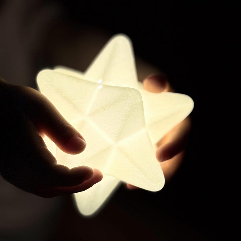 May include: A glowing, star-shaped night light held against a dark background. The white light emits from the star's geometric design. The star is held in two hands, with the fingers visible.