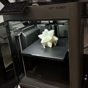 May include: A 3D printer with a geometric star-shaped object on the print bed. The printer is gray with the text "Bambu Lab X2D" on the side. The object is white and has a complex, multi-faceted design.