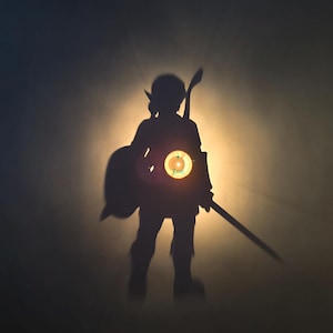 May include: A dark silhouette of a humanoid figure standing against a bright circular light source. The figure wears a pointed cap, carries a shield on one arm, and holds a sword in the other hand. A small, circular emblem with text is visible.