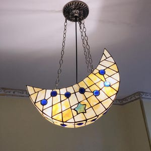 Può includere: Un lampadario Tiffany a forma di mezzaluna. La lampada presenta un design a mosaico con vetro giallo e bianco, accentuato da cerchi blu e una stella verde-blu. È appesa a un supporto a soffitto decorativo tramite catene.