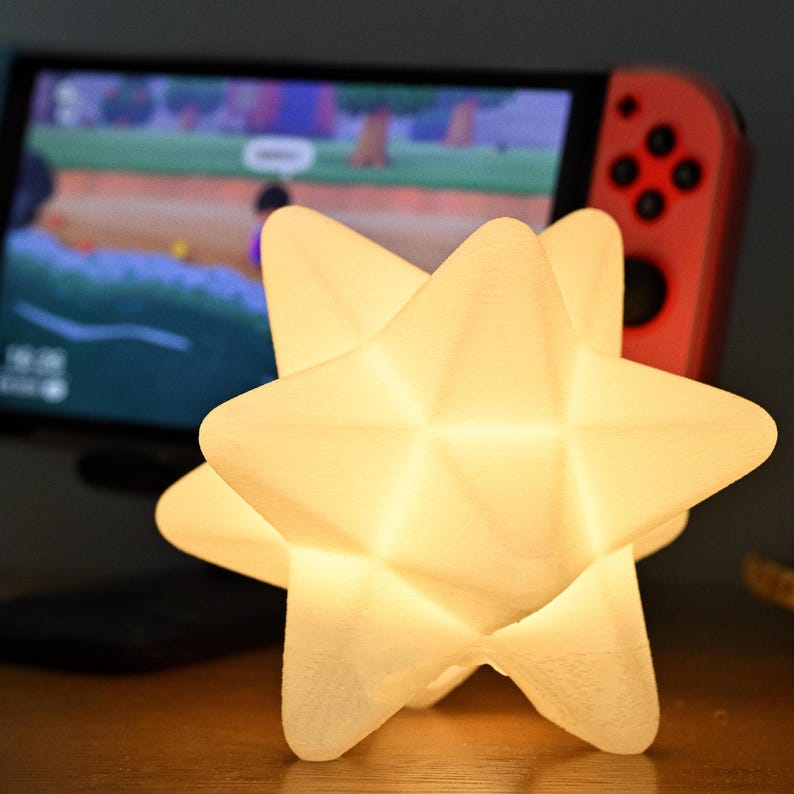 May include: A glowing, star-shaped lamp with a geometric design. The lamp emits a soft, warm light. A Nintendo Switch console is visible in the background. The lamp is on a wooden surface.
