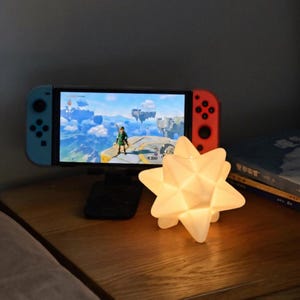 May include: A Nintendo Switch console with a game displayed on the screen, supported by a black stand. A glowing white star-shaped lamp sits in front of the console on a wooden surface. The console has blue and red controllers.
