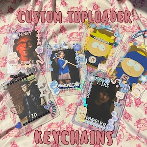 May include: Custom toploader keychains featuring various images, including characters and celebrities. The clear plastic holders are decorated with beads, stars, and bows. Text on the keychains includes names like Henry, Veronica, JD, and Mike.
