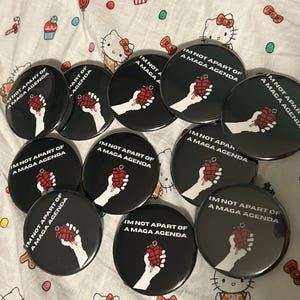 Op de afbeelding: Een verzameling zwarte ronde buttons met de tekst "I'M NOT APART OF A MAGA AGENDA". Elke button toont een witte hand die een rode granaat vasthoudt. De buttons liggen op een wit oppervlak met een patroon.