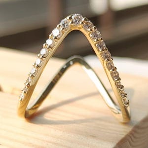 Pode incluir: Um anel dourado com design em forma de V, cravejado com pequenas pedras preciosas transparentes. O anel é exibido em um bloco de madeira clara, mostrando seus detalhes delicados. O design do anel é moderno e elegante.