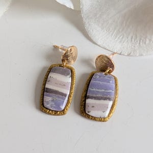 Op de afbeelding: Een paar goudkleurige oorstekers met rechthoekige hangers met marmer-effect in tinten lavendel, wit en grijs. De oorbellen hebben een getextureerde gouden rand en een kleine, ronde gouden stud.