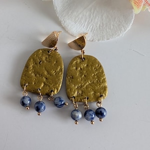 May include: Gold-toned earrings with textured, irregular shapes and small, dangling blue and white beads. The earrings have a bird-like design at the top and are suspended from gold-colored posts.