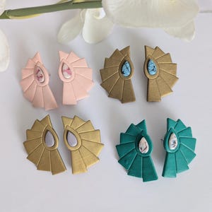 May include: Four pairs of handmade earrings in various colors. The earrings feature a fan-like design with a teardrop-shaped stone in the center. Colors include pink, gold, teal, and a gold-tone with a gray stone. The earrings are displayed on a white surface.