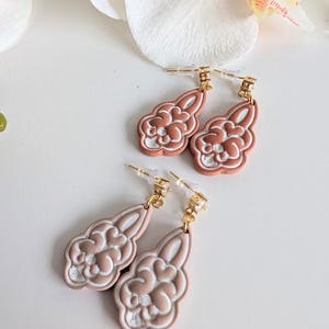 Könnte beinhalten: Zwei Paar Ohrhänger mit wolkenförmigem Design. Die Ohrringe sind hellbraun mit weißen Akzenten und goldfarbenen Stiften. Sie werden auf einer weißen Oberfläche mit einer weißen Blume im Hintergrund präsentiert. Ein dekoratives Accessoire.