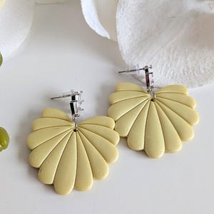 Puede incluir: Un par de pendientes amarillo pálido con un diseño en forma de abanico. Los pendientes tienen un poste y un respaldo plateados. Los pendientes están sobre una superficie blanca con una flor blanca en el fondo. Un accesorio único.