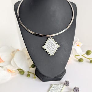 May include: A silver choker necklace with a geometric pendant and matching earrings. The pendant and earrings have a white and light green diamond-shaped design with a detailed pattern. The jewellery is displayed on a black mannequin head.