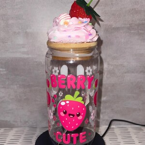 Puede incluir: Un tarro de cristal transparente con tapa de bambú, decorado con un diseño de fresa rosa y las palabras "BERRY CUTE". El tarro está cubierto con glaseado rosa, chispas y una fresa roja. El tarro se encuentra sobre una base giratoria negra.