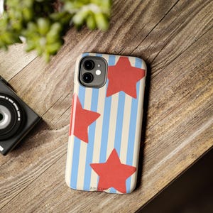 May include: A mobile phone case featuring a pattern of vertical light blue and cream stripes overlaid with three large, solid red stars. The case has a cutout for a dual-lens camera system on the top left corner and the text JUJUSTUDIO at the bottom.
