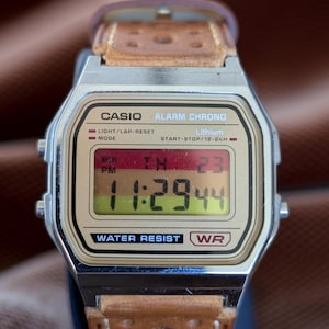 Puede incluir: Un reloj digital Casio con correa de cuero marrón. La pantalla muestra la hora 23:29:44. El reloj tiene una caja plateada y muestra el texto "CASIO ALARM CHRONO" y "WATER RESIST".