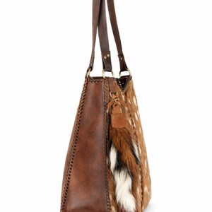 May include: A brown shoulder bag featuring a smooth leather side panel with decorative whipstitching, a textured animal print front panel, two long leather straps, and a hanging tassel ornament made of brown, white, and black fur and leather.