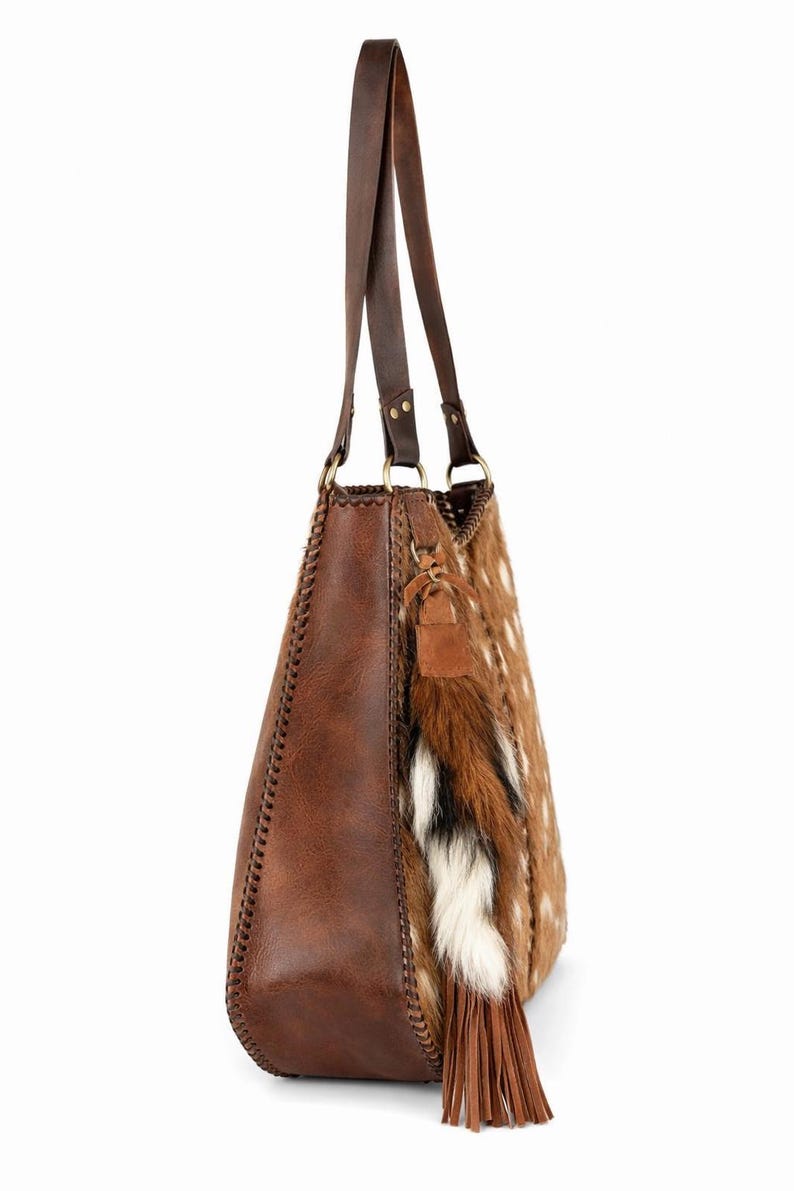May include: A brown shoulder bag featuring a smooth leather side panel with decorative whipstitching, a textured animal print front panel, two long leather straps, and a hanging tassel ornament made of brown, white, and black fur and leather.