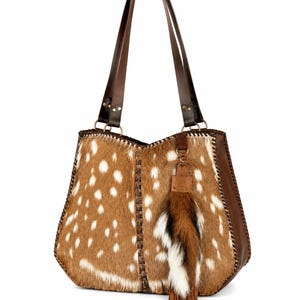 May include: A tote bag crafted from brown and white spotted animal hide featuring dark brown leather side panels and shoulder straps. The bag includes a decorative hanging tassel made of brown and white fur with leather fringe accents.