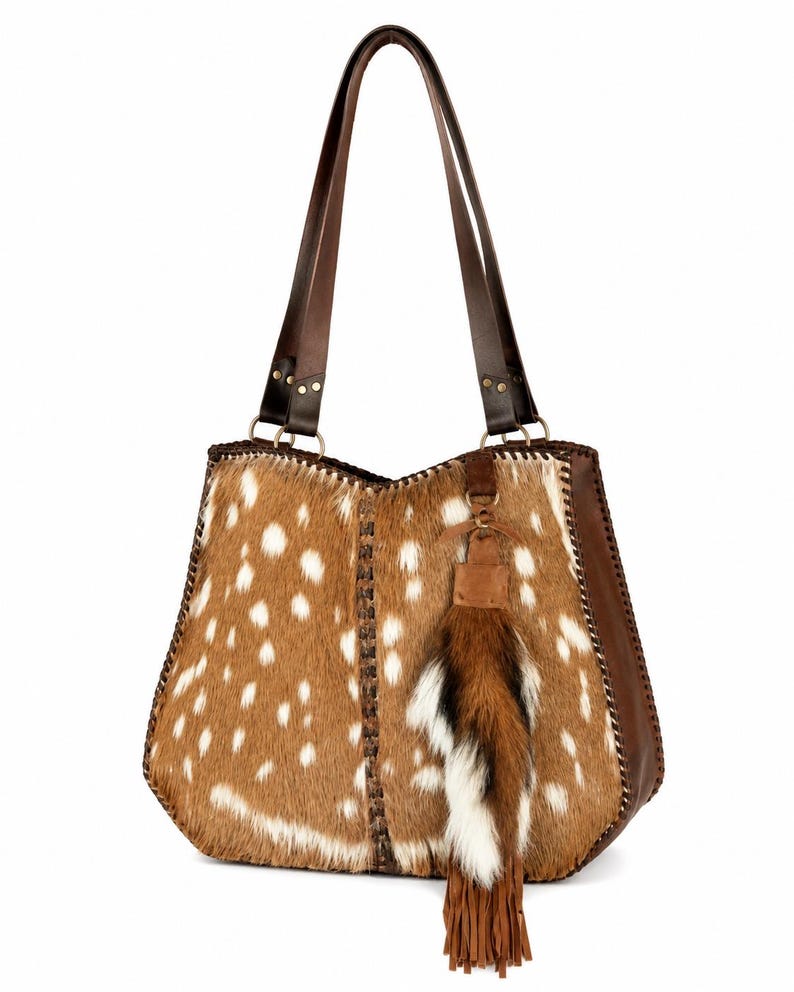 May include: A tote bag crafted from brown and white spotted animal hide featuring dark brown leather side panels and shoulder straps. The bag includes a decorative hanging tassel made of brown and white fur with leather fringe accents.